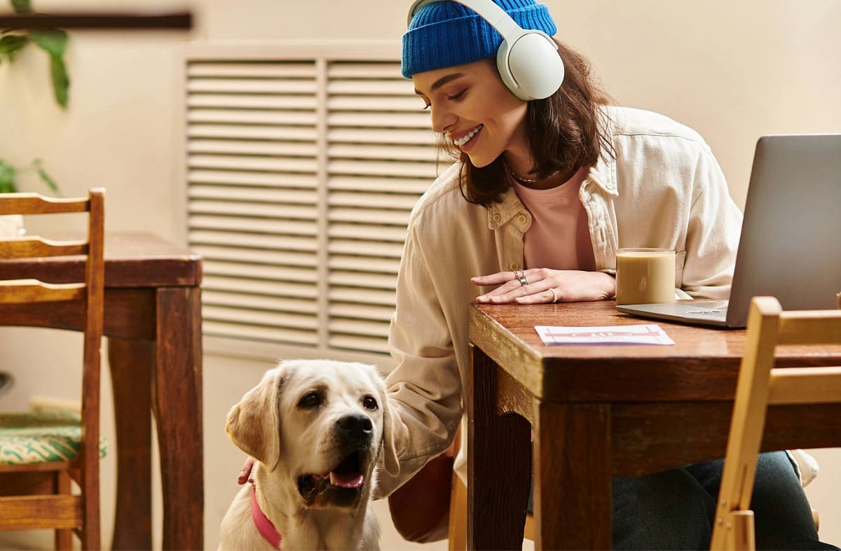 female sitting at table with coffee and laptop listening to headphones petting a Labrador Retriever